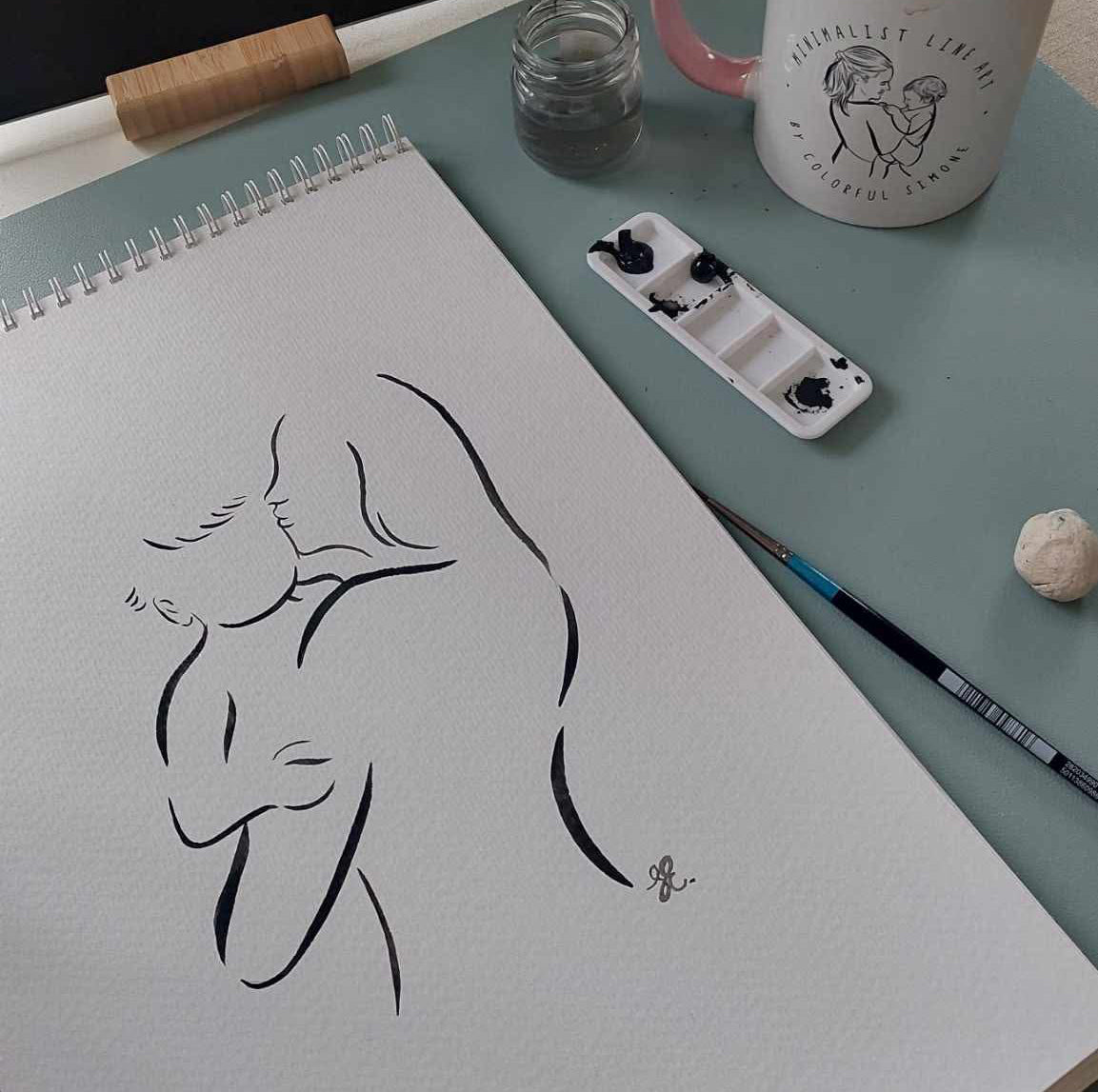 Artistic sketch of a nude figure on a drawing pad with a mug and watercolor set in the background.